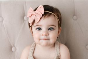 Cute baby girl with green eyes wearing a pink bow headband against a tufted beige background.