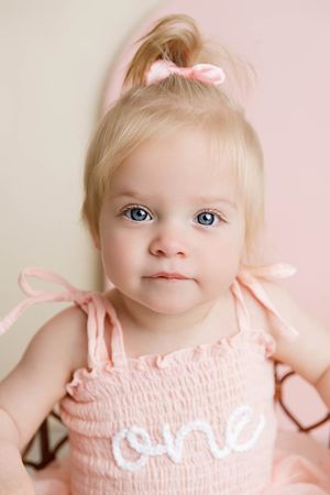 Baby's 1st birthday portrait of blue-eyed blonde baby girl in pink smocked dress with one written on it.