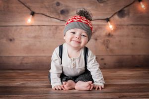 Baby boy's Menifee 6 month photoshoot, smiling in holiday knit pom pom hat with suspenders and string lights
