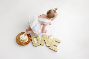Baby girl in white lace dress reaching toward birthday cake beside gold ONE letters.