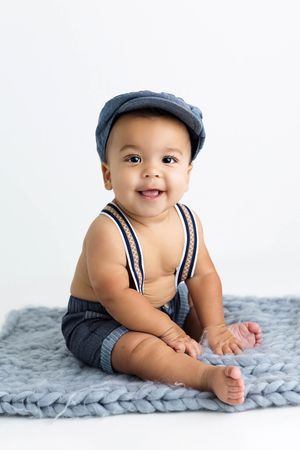 Smiling baby boy in blue cap and suspenders sitting on chunky knit blue blanket.