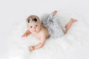 6 month photoshoot of baby girl lying on white fur rug wearing a gray tutu skirt and bow headband.