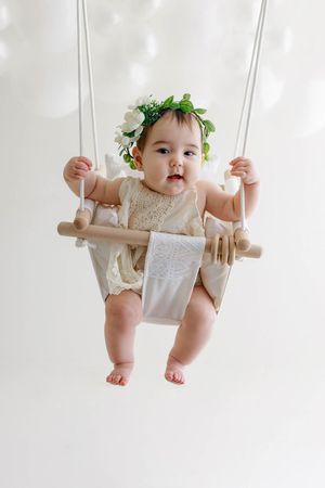 Adorable baby in floral crown sitting in infant swing surrounded by white balloons.