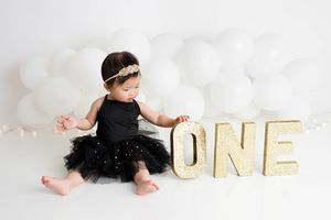 Baby girl in black tutu dress sitting next to gold glitter ONE letters with white balloons.
