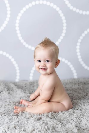 Sitter session in Menifee: Happy baby with mohawk hairstyle sitting on gray fluffy carpet smiling at camera.