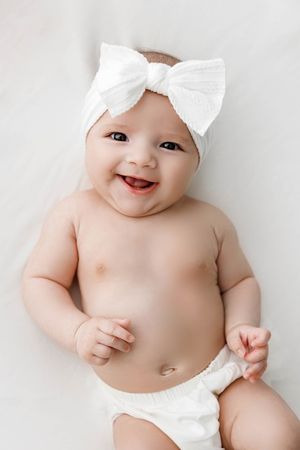 Happy smiling baby girl wearing white bow headband lying on white background.