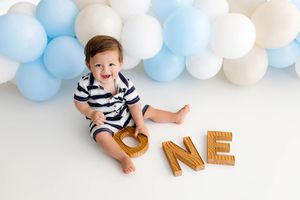 Baby boy in striped outfit sits smiling among blue and white balloons with wooden ONE letters for first birthday.