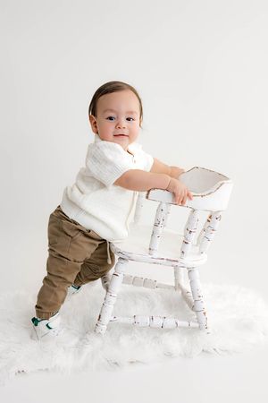A baby boy in a white sweater and khaki pants stands beside a distressed white wooden chair on a white fur rug.