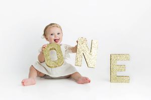 Cute baby girl in white dress holding gold glitter ONE letters celebrating her first birthday.