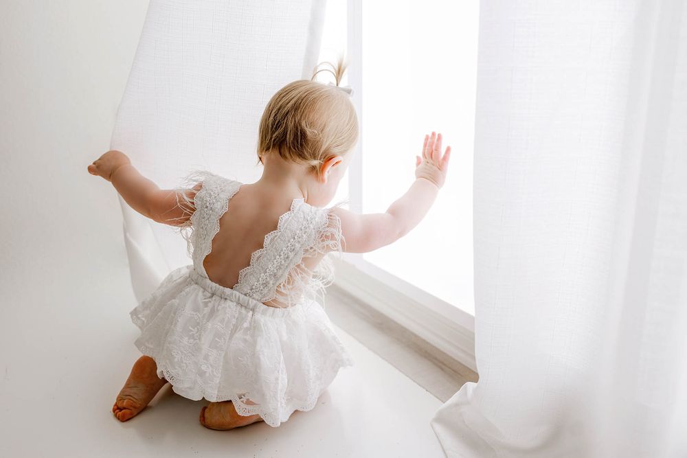 Toddler in white lace romper reaching toward bright window with sheer curtains.
