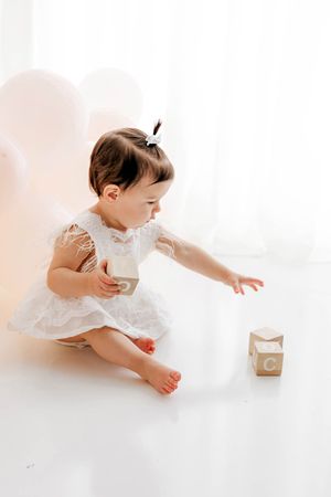 Baby girl in white lace dress reaching for wooden blocks spelling out ONE for 1st birthday photos on bright white backdrop.
