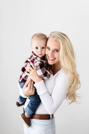 Mom and baby photoshoot in Menifee, smiling mom holding baby boy in plaid shirt against white background.