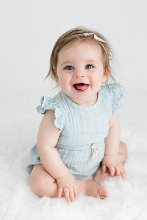 Smiling baby girl in light blue ruffle romper with white bow headband sitting on fluffy white rug.