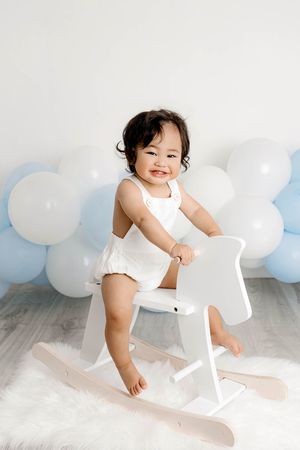 Baby boy's 1st birthday photoshoot in Menifee, wearing white romper riding white rocking horse with blue balloon backdrop.