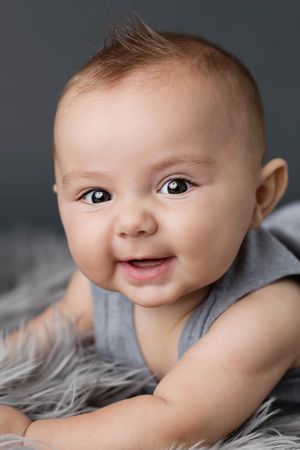 Smiling baby boy in gray outfit lying on fluffy gray rug against dark gray background.