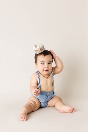 Cute baby boy wearing crown and suspenders sitting on cream background for first birthday photo.