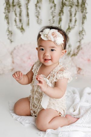 Cute baby girl in lace romper with pearl necklaces and floral headband sitting against floral backdrop.