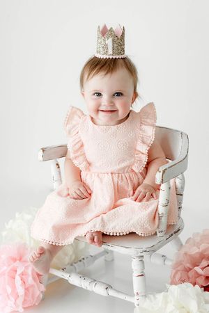 Happy baby girl in pink ruffle dress with gold glitter crown sitting in vintage white chair.