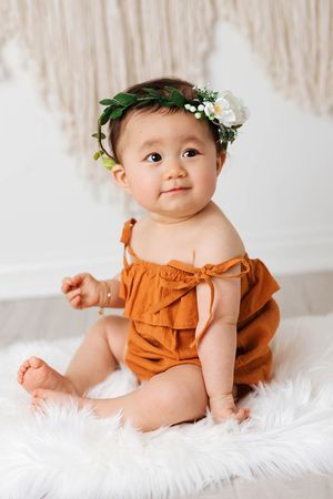 Sweet baby girl wearing orange off-shoulder romper and floral crown sitting on white fur rug.