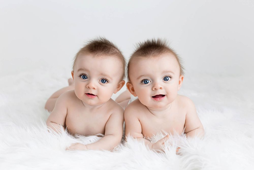 Adorable twin babies with blue eyes lying side by side on white fluffy blanket.