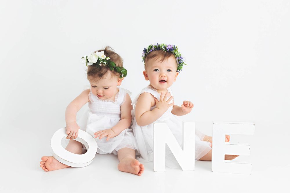 Twin baby girls in white rompers with flower crowns sitting beside large ONE letters for first birthday photoshoot in Murrieta.