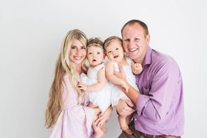 Menifee family and child photographer: Smiling family of four with parents holding twin baby girls in white dresses against white backdrop.