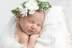 Sleeping newborn baby girl wearing a delicate white floral crown on white blanket.