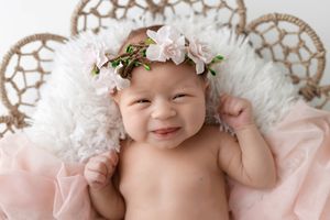 Happy newborn baby girl wearing pink floral crown lying on fluffy white rug with pink fabric.