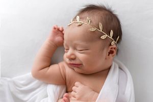 Sleeping newborn baby girl smiling with gold leaf headband wrapped in white swaddle.