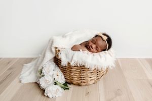 Newborn baby sleeping in a wicker basket with white fur and fresh white peony flowers.