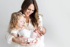 Smiling mother in white dress holding newborn baby while toddler daughter looks on lovingly.