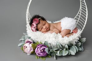 Sleeping newborn baby girl in white fluffy basket with pink flowers and green leaves on gray background.