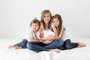 Three sisters sitting together on white bed smiling while oldest holds newborn baby sister.