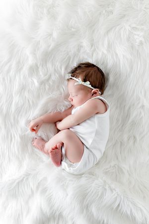 Sleeping newborn baby girl in white romper and floral headband on fluffy white rug.
