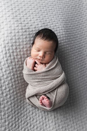 Peaceful newborn baby swaddled in gray knit wrap on textured gray blanket.