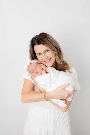 Smiling mother in white dress holds sleeping newborn with pink bow headband.