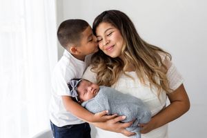 Mother holds swaddled newborn while young son gives her a loving kiss on cheek.