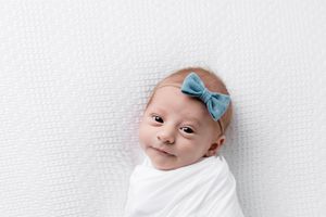 Portrait of newborn baby girl swaddled in white with blue bow headband.