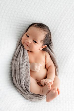 Alert newborn baby wrapped in gray knit swaddle smiling on white blanket.