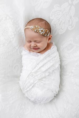 Smiling newborn baby swaddled in white lace wrap with jeweled gold headband.