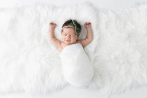 Newborn baby wrapped in white swaddle on fluffy white fur rug with green floral headband.