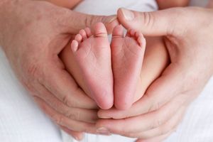 Dads hands forming heart shape around tiny newborn baby feet on white background.