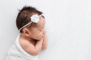 Sleeping newborn baby in white swaddle with white rose headband on white background.