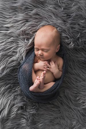 Sleeping newborn baby wrapped in blue knit wrap on gray fluffy fur rug.