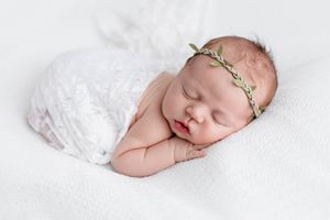 Newborn baby sleeping swaddled in white lace wrap with a green leaf headband.