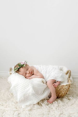 Newborn baby girl sleeping in a wicker basket with a floral crown on white fur.