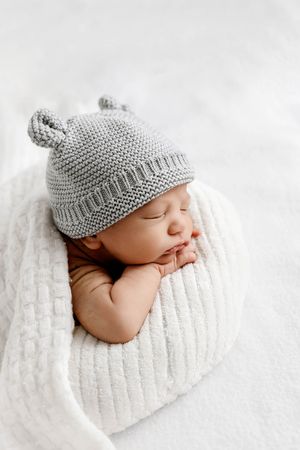 Cute newborn baby sleeping in a white blanket wearing a gray knit bear hat.