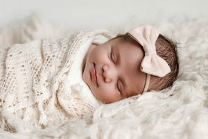 Smiling sleeping newborn baby wrapped in a cream knit blanket with a pink bow headband.