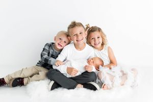 Three smiling siblings pose together holding their newborn baby brother on white background.
