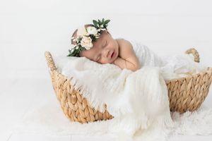 Sleeping newborn baby with floral crown resting in wicker basket with white fur blanket.